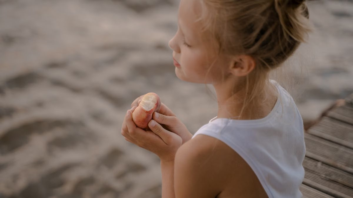 Blond girl enjoying a peach on a pier by the water. Serene summer day.